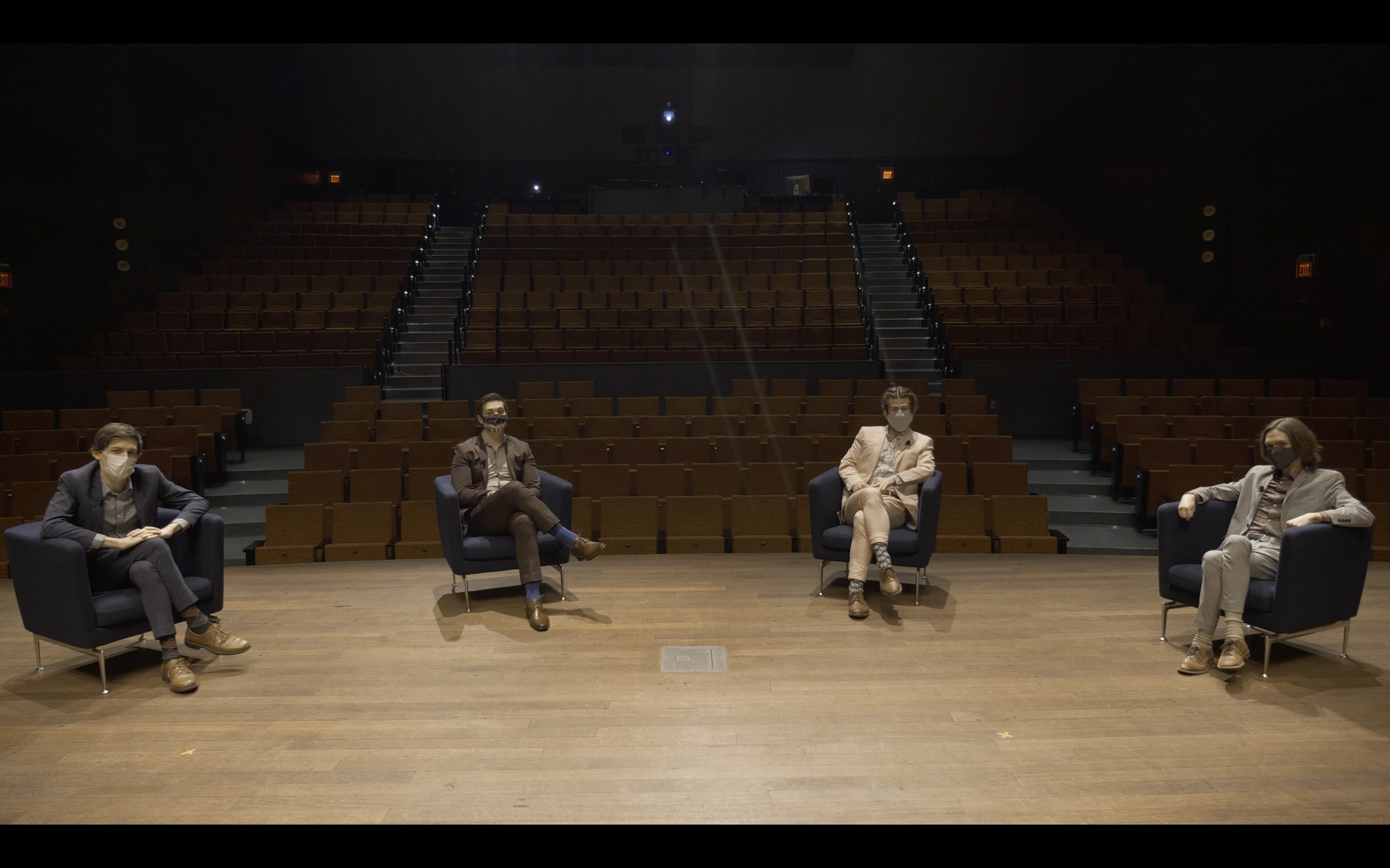 The four members of ~Nois sit in chairs spaced apart, wearing suits and facemasks, in the Logan Center Performance Hall