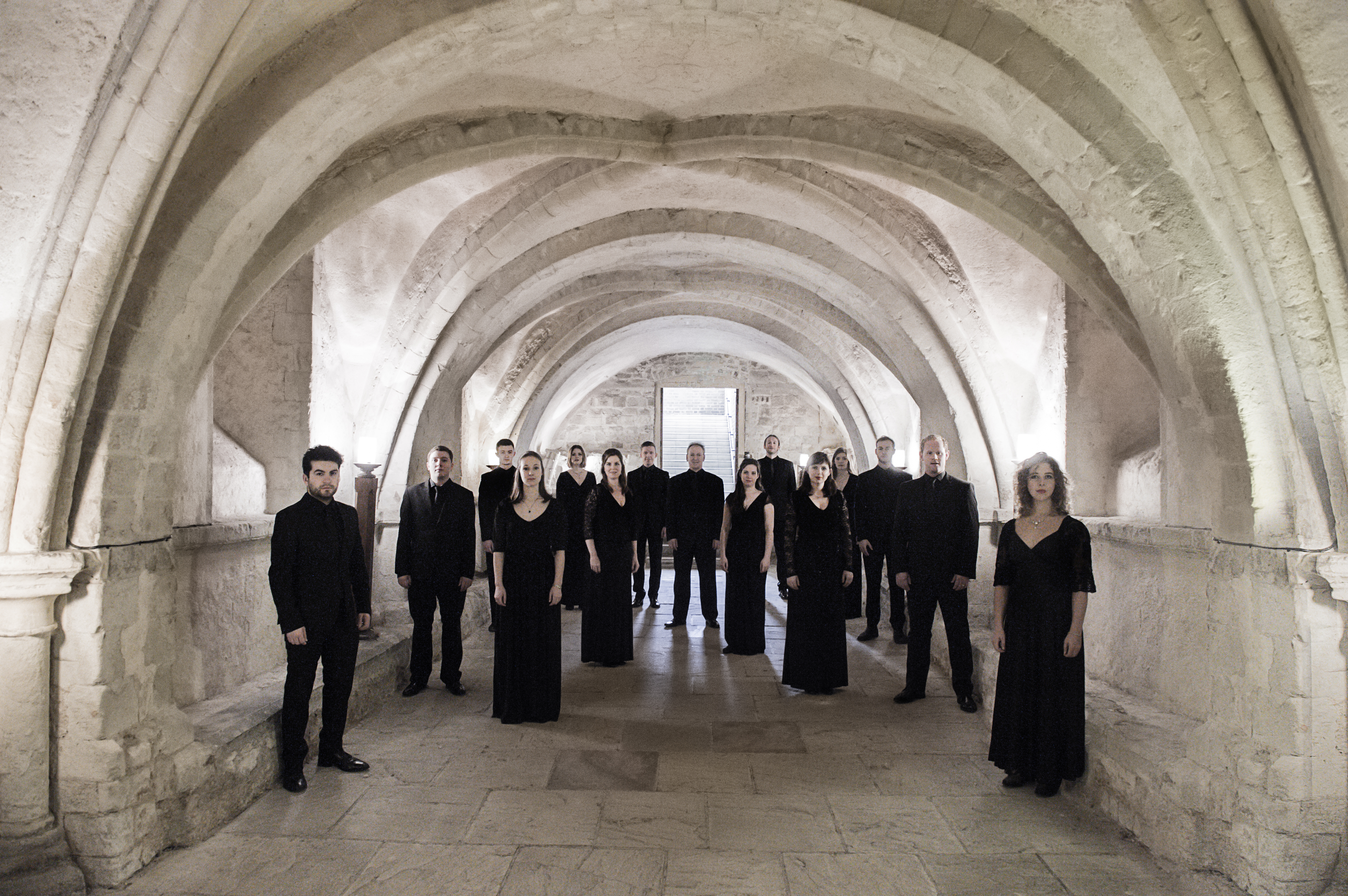 The members of the choir Tenebrae wearing black stand in a brightly lit stone vaulted hallway