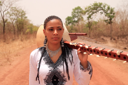 A woman walking on a path in nature with a stringed instrument over her shoulder.
