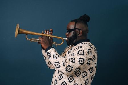 A man in a patterned shirt plays the trumpet in front of a dark blue background
