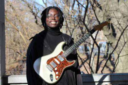 A man in a black jacket stands outside, smiling and holding a white and red guitar