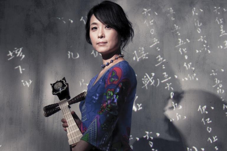 Wu Man holds a pipa in front of a slate wall with Chinese characters written in chalk