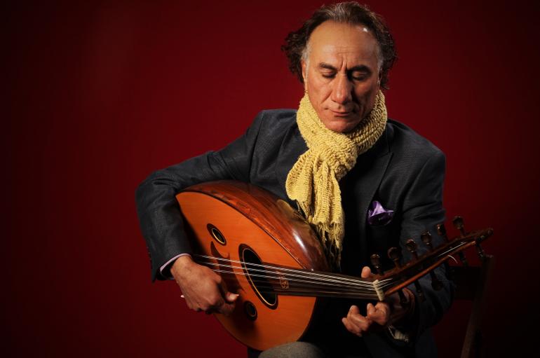 Rahim AlHaj playing oud, wearing a yellow scarf in front of a red background