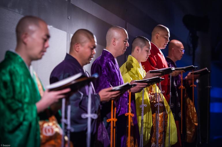 Six monks wearing brightly colored silk robes is shown chanting while holding chant books