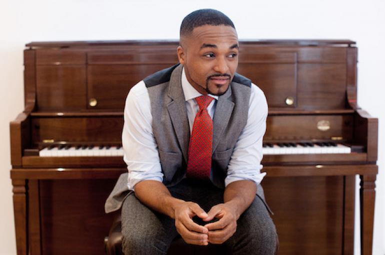Sullivan Fortner sitting in front of a brown upright piano, wearing a grey vest and pants, white shirt, and red tie