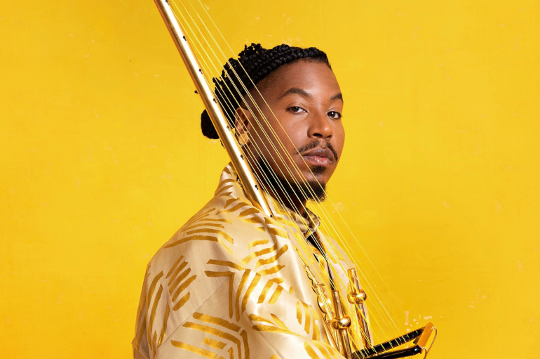 A man posing in front of a yellow background and holding a stringed instrument