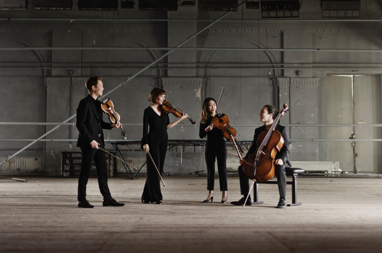 A candid photo of a string quartet against an industrial background with instruments in hand