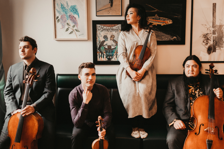 Four people pose on a sofa with string instruments in hand
