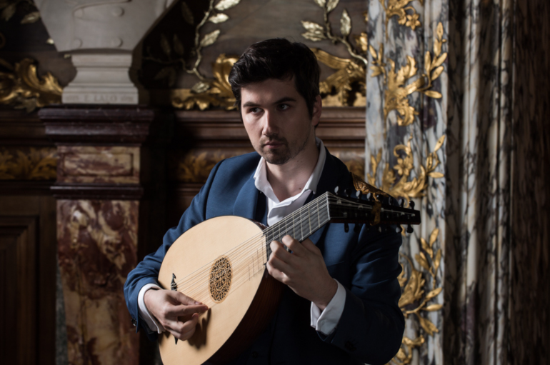A man stands against an ornate background, playing the lute. 
