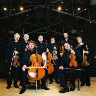 The Academy of St. Martin in the Fields Chamber Ensemble stands together dressed all in black with their instruments