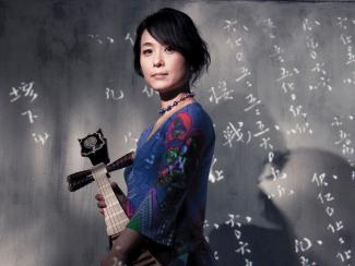 Wu Man stands holding a pipa in front of a slate wall with Chinese characters written in chalk