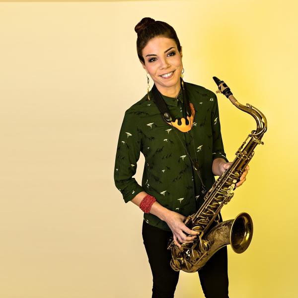 Melissa Aldana holding saxophone and standing in front of yellow background