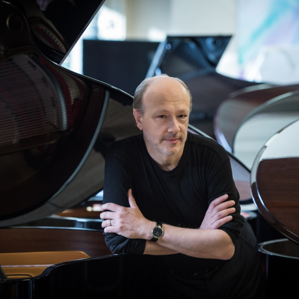 Marc-André Hamelin wearing a black sweater and leaning on a gran piano when many open grand pianos behind him
