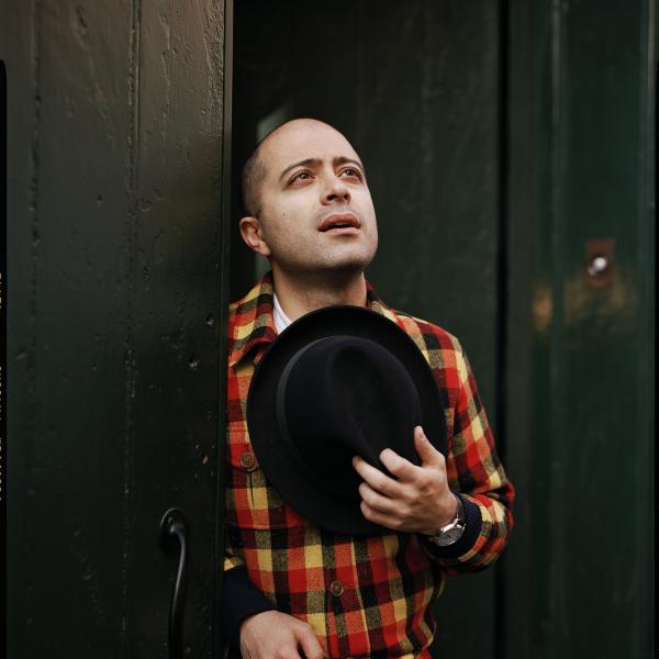Mahan Esfahani shown wearing a red, brown, and yellow plaid jacket and holding brown hat to his chest
