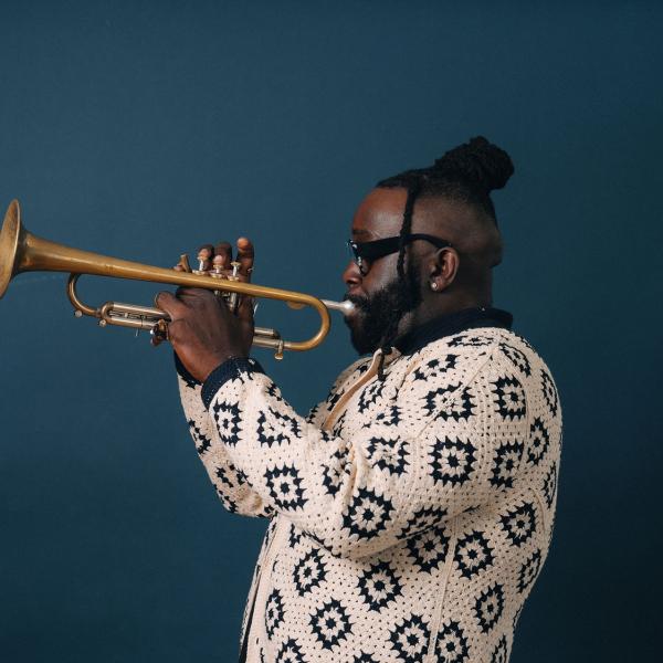 A man in a patterned shirt plays the trumpet in front of a dark blue background