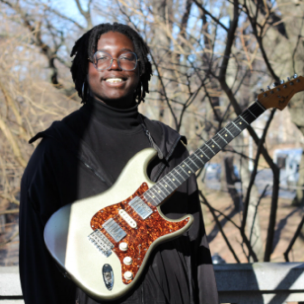 A man in a black jacket stands outside, smiling and holding a white and red guitar